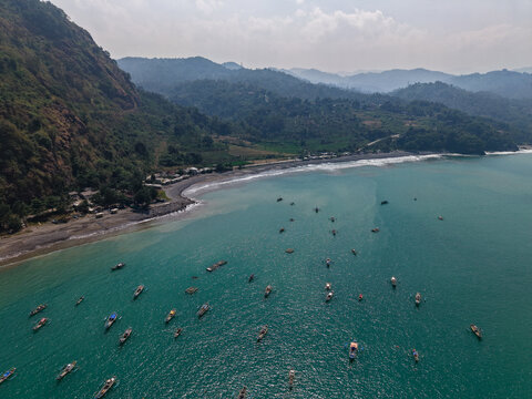 Aerial view of Pantai Cikembang with numerous small fishing boats anchored in the turquoise bay, surrounded by lush green hills in Kecamatan Cisolok, Jawa Barat, Indonesia.