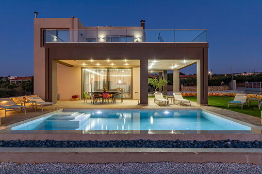 View of a modern villa with a swimming pool at dusk, featuring an illuminated interior, outdoor dining area, lounge chairs, and a manicured lawn in Chania, Greece.