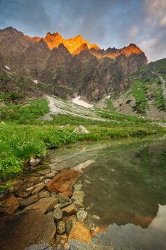 View of Zelene Kacacie pleso in Litvorova dolina under the golden-lit peaks of Ganok mountain with rocky shores and lush green vegetation in Tatranska Javorina, Presovsky kraj, Slovakia.