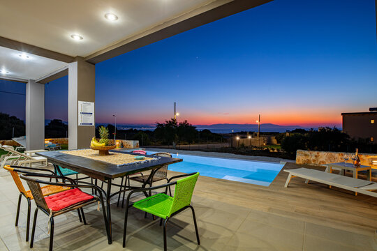 View of a modern patio with a dining table and colorful chairs overlooking a swimming pool and the sea at sunset in Chania, Greece.