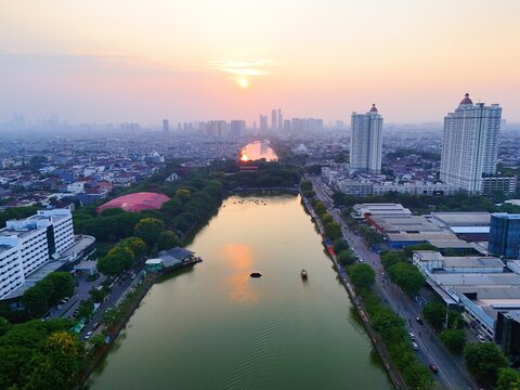 Aerial view of Danau Sunter at sunset with the surrounding urban skyline, lush trees, and calm water reflecting the golden light in Kecamatan Tanjung Priok, Daerah Khusus Ibukota, Indonesia.