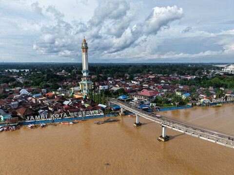 Aerial view of Jembatan Gentala Arasy and its iconic tower spanning the brown Batang Hari river with dense urban rooftops under a cloudy sky in Kecamatan Jambi Timur, Jambi, Indonesia.