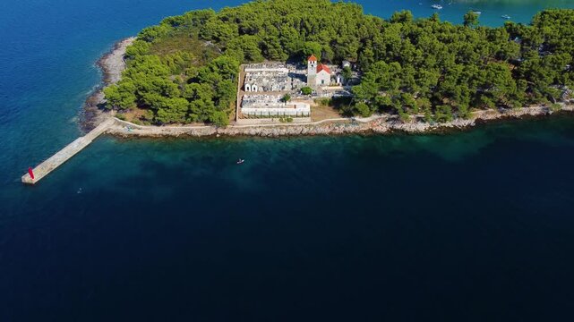 Aerial view of Jelsa Cemetery surrounded by dense pine forest and turquoise Adriatic Sea waters in Jelsa, Split-Dalmatia County, Croatia.