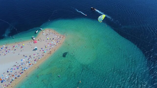 Aerial view of Golden Horn Beach with people on the sand, boats in the turquoise water, and a parasailer in the air in Bol, Split-Dalmatia County, Croatia.