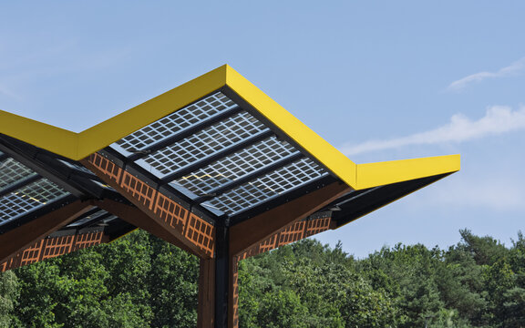 View of the Fastned Charging Station with its distinctive yellow roof and integrated solar panels, set against a clear blue sky and lush green trees in Arnhem, Gelderland, Netherlands.