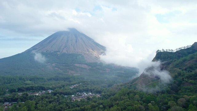 Aerial view of Mount Inerie volcano with lush green slopes, clouds drifting around the peak, and a small village in the valley below in Inerie, East Nusa Tenggara, Indonesia.