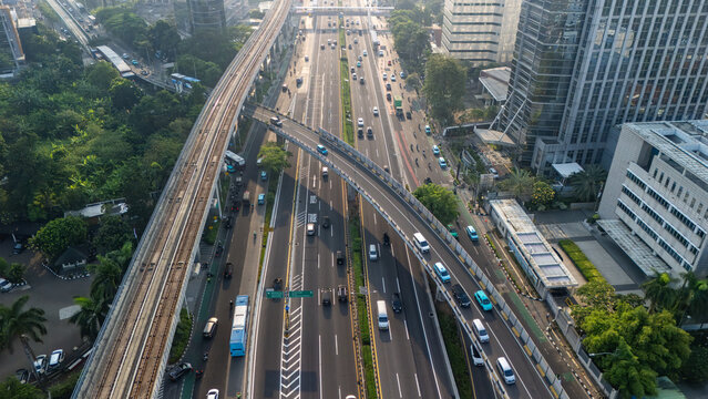 Aerial view of a busy multi-lane highway with elevated train tracks, modern office buildings, and dense urban traffic, Jakarta, Indonesia.