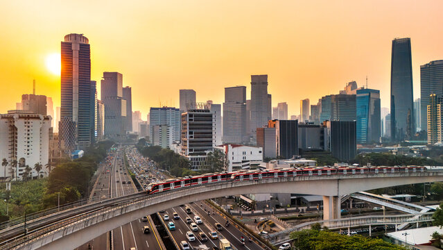 Aerial view of the modern Jakarta skyline at sunset, featuring an elevated train moving along tracks above a busy highway with dense traffic, Jakarta, Indonesia.