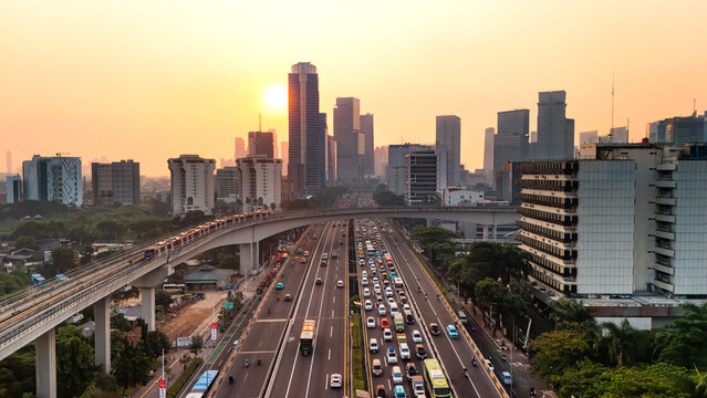 Aerial view of the Jakarta skyline at sunset with a busy highway and elevated train track, surrounded by modern skyscrapers and lush greenery in Jakarta, Indonesia.