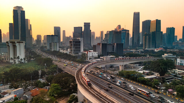 Aerial view of the Jakarta skyline featuring a modern train on an elevated track above a busy highway with skyscrapers and golden sunset light in Jakarta, Indonesia.