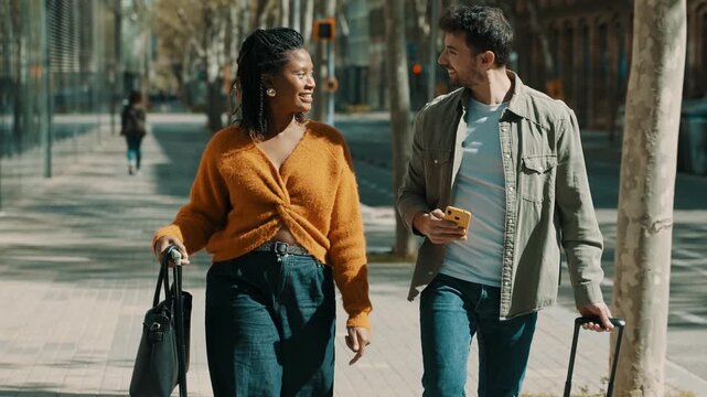 Smiling tourist couple walking with luggage along an urban city sidewalk during vacation travel.