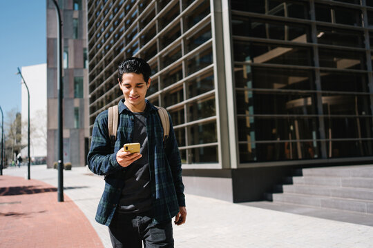 Young hispanic student walking using mobile phone app in the city, in modern business area