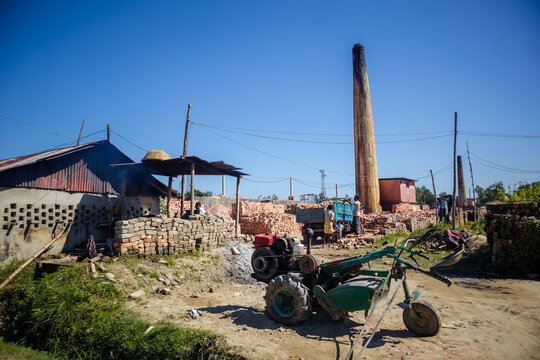Feni, Chittagong Division, Bangladesh - 19 November 2024: View of a brick kiln with a tall chimney, workers stacking bricks, and a two-wheeled tractor in a rural industrial setting.