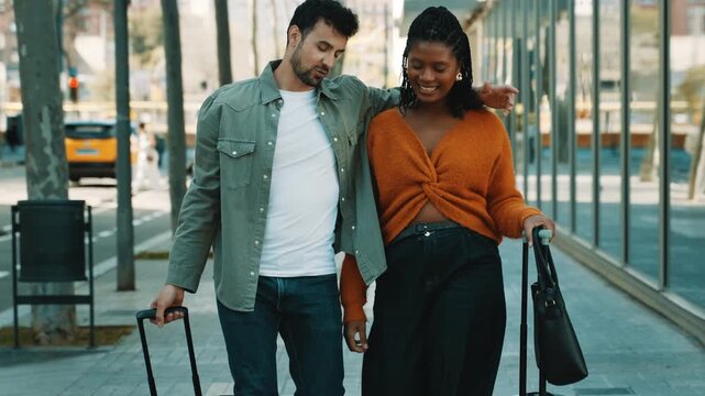 Multicultural tourist couple walking with luggage along an urban city sidewalk during vacation travel