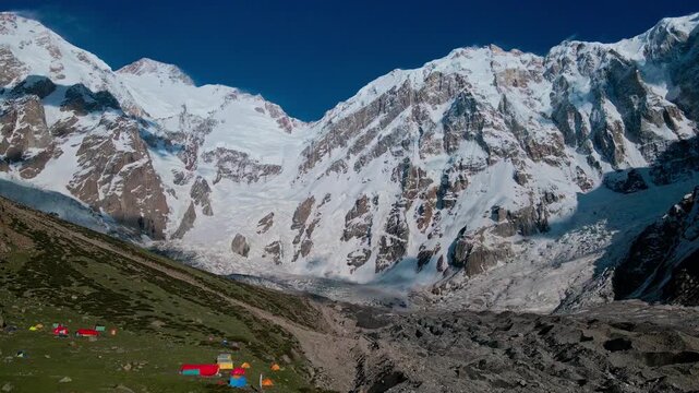 Aerial view of the massive snow-covered Diamir face of Nanga Parbat with a base camp featuring colorful tents on the rocky glacier in Diamir, Gilgit Baltistan, Pakistan.