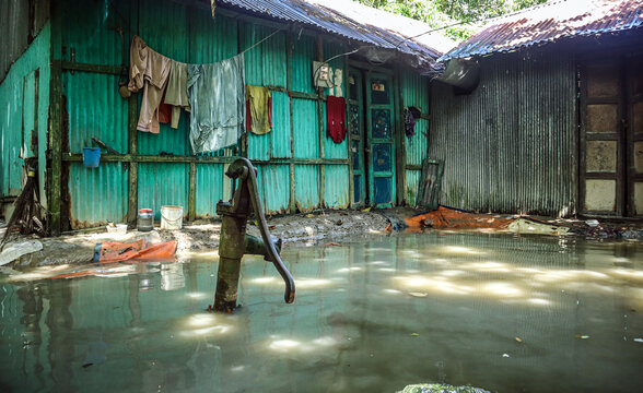 Noakhali, Bangladesh - 02 September 2024: View of a flooded residential area with a manual water pump standing in murky water in front of houses with corrugated metal walls and hanging laundry.