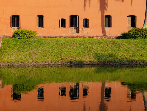 Sonargaon, Bangladesh - 19 November 2025: View of the historic orange building in Panam City, featuring arched windows with iron bars, a green grass bank, and its reflection in water.