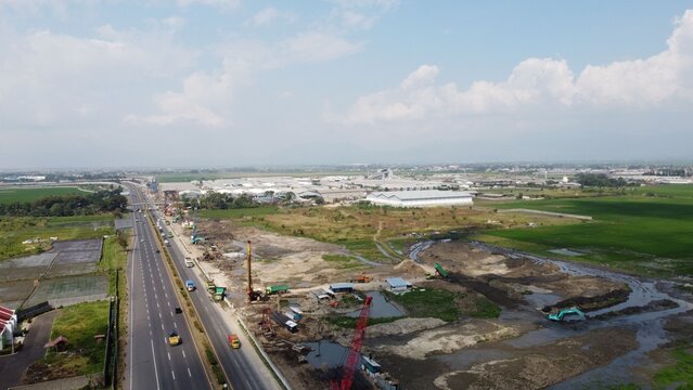 Aerial view of the Kertajati highway construction site with heavy machinery, cranes, and earthworks alongside a multi-lane road near Bandung, West Java, Indonesia.