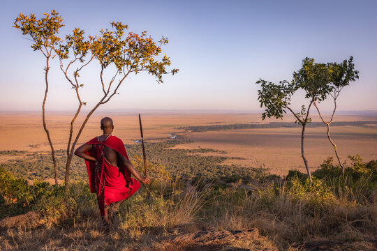View of a Maasai warrior in traditional red clothing holding a spear while standing on a hill overlooking the vast savanna landscape in Kenya.