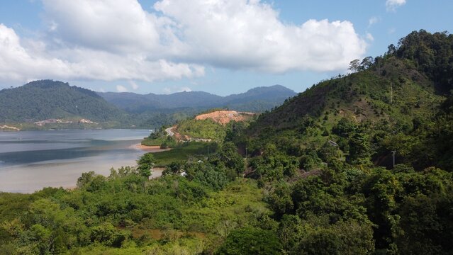 Aerial view of the Mandeh coastal area featuring lush green hills, a winding road along the bay, and calm waters in Koto XI Tarusan, West Sumatra, Indonesia.