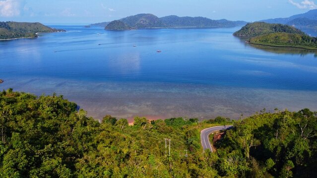 Aerial view of Mandeh, featuring a winding coastal road through lush tropical forest overlooking the calm blue waters of the bay in Koto XI Tarusan, West Sumatra, Indonesia.