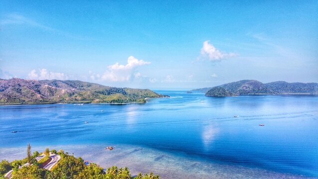 Aerial view of Mandeh coastal area with lush green hills, winding roads, and clear blue waters in Koto XI Tarusan, West Sumatra, Indonesia.