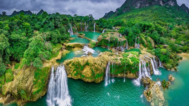 View of Ban Gioc Waterfalls cascading over lush green rocky tiers into turquoise pools, surrounded by dense forest and limestone mountains in Dam Thuy, Cao Bang, Vietnam.