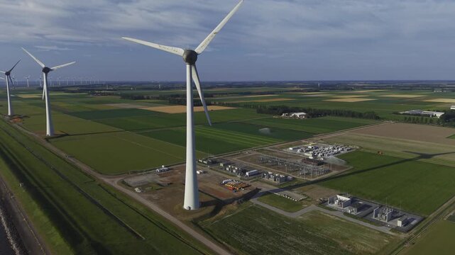 Aerial view of wind turbines standing in a vast green agricultural landscape with a power substation nearby under a clear sky in Espel, Flevoland, Netherlands.