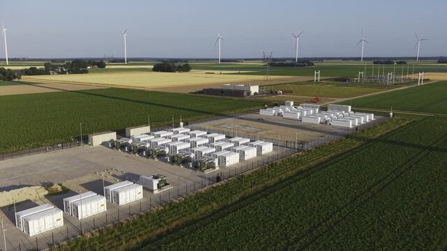 Aerial view of a large-scale battery energy storage system with rows of white containers situated amidst agricultural fields and wind turbines in Dronten, Flevoland, Netherlands.