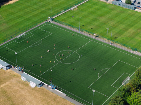 Aerial view of a football pitch with players in action, surrounded by green fields and floodlights in Studley, England, United Kingdom.