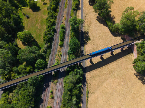 Aerial view of Edstone Aqueduct carrying a narrow boat over the railway tracks and road, surrounded by lush green trees and dry golden fields in Wootton Wawen, England, United Kingdom.