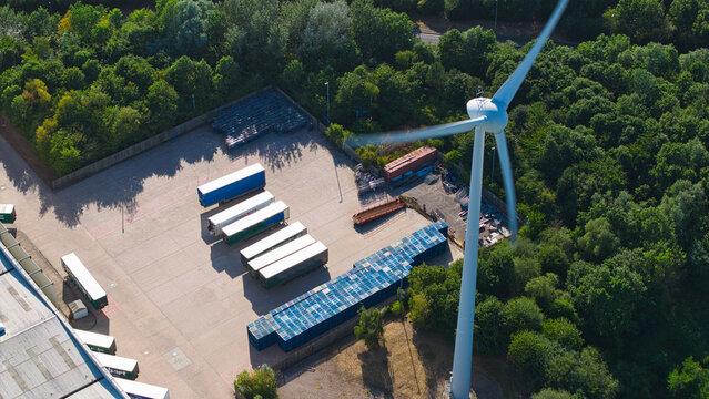 Aerial view of a wind turbine standing next to a logistics yard filled with parked HGV trailers and shipping containers in Crick, England, United Kingdom.