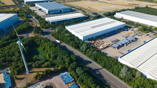 Aerial view of Daventry International Rail Freight Terminal featuring large warehouses, distribution centers, and parked HGVs near a wind turbine in Kilsby, England, United Kingdom.