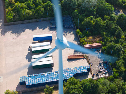 Aerial view of a wind turbine with rotating blades positioned above a logistics yard filled with parked semi-trailers and shipping containers in Crick, England, United Kingdom.