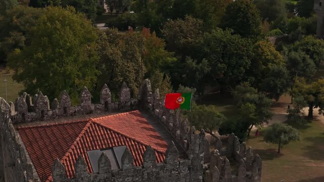 Aerial view of Guimaraes Castle with its stone battlements and the Portuguese flag flying over the tiled roof, surrounded by lush parkland in Guimaraes, Braga, Portugal.