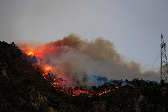 Aerial view of a forest fire burning on a hillside in the Susa Valley with thick smoke and flames rising near a power line tower in Chianocco, Piedmont, Italy.