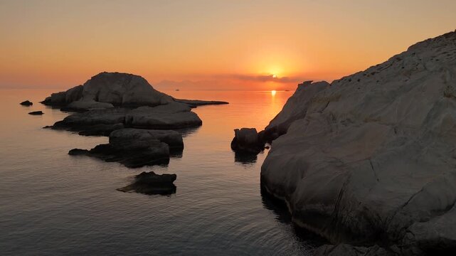 Peaceful sunrise over the calm Aegean Sea at Mandrakia on the Greek island of Milos. The stationary camera captures warm orange and red hues reflecting on tranquil water and white coastal rocks.