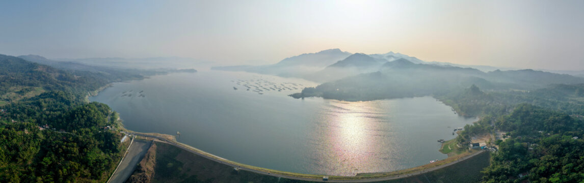 Aerial view of Wadaslintang Reservoir with calm water, floating fish cages, surrounding lush green hills, and misty mountains, Wadaslintang, Central Java, Indonesia.