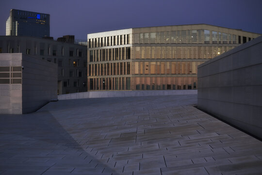 Oslo, Norway - 16 July 2026: View of the iconic marble-clad roof of the Oslo Opera House at dusk, with modern office buildings in the background under a deep blue sky.
