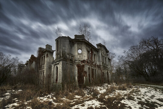 Poland - 21 February 2017: View of an abandoned, ruined stone mansion overgrown with vegetation under a dramatic, cloudy sky in winter.