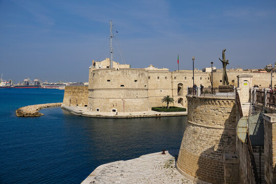 View of the historic Castello Aragonese fortress on the waterfront, featuring stone walls, a tower, and a bronze sculpture on the foreground terrace in Taranto, Apulia, Italy.