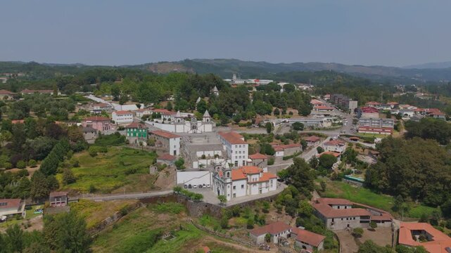 Aerial view of Santuario de Nossa Senhora de Porto de Ave surrounded by lush green hills, stone buildings, and winding roads in the rural landscape of Taide, Braga, Portugal.