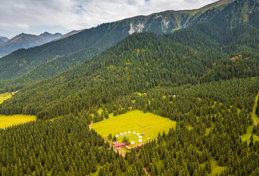Aerial view of Kok Jaiyk valley featuring a cluster of yurts in a lush green meadow surrounded by dense coniferous forests and mountains in Dzhetyoguz, Issyk-Kul Region, Kyrgyzstan.