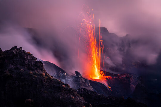 View of Stromboli volcano erupting with glowing lava fountains and volcanic ash plumes rising into the night sky, Lipari, Sicily, Italy.