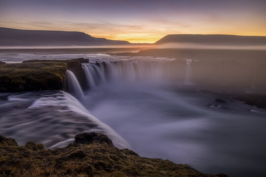 View of the majestic Godafoss waterfall at sunset with misty water flowing over rocky cliffs in Iceland.