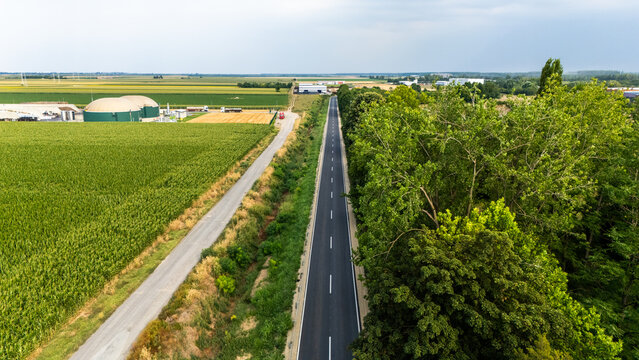 Aerial view of a straight road passing through agricultural fields and a forest with industrial biogas plant domes in the background in Serbia.