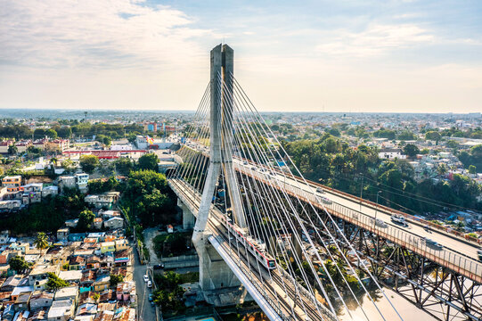 Aerial view of the Puente Juan Bosch cable-stayed bridge crossing the Ozama River with a metro train passing by and dense urban neighborhoods in Santo Domingo, Dominican Republic.