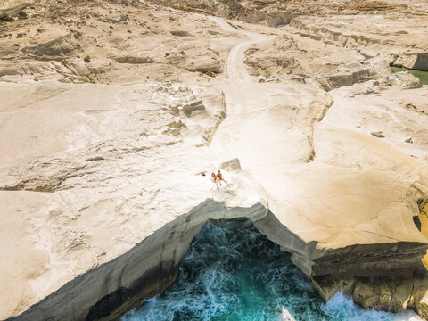 Aerial view of Sarakiniko Beach with its unique white volcanic rock formations and turquoise sea, featuring a couple sitting on the edge of a cliff in Milos, Greece.
