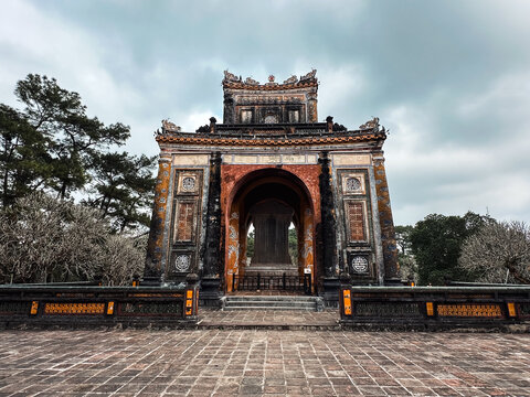 Hue, Vietnam - 06 February 2026: View of the historic stele pavilion at the Tu Duc Tomb in Hue, featuring intricate traditional architecture, weathered stone walls, and a cloudy sky.