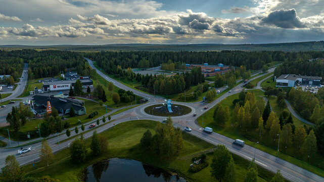 Kuusamo, North Ostrobothnia, Finland - 10 September 2025: Aerial view of a roundabout surrounded by lush coniferous forests, commercial buildings, and a small pond under a dramatic cloudy sky.